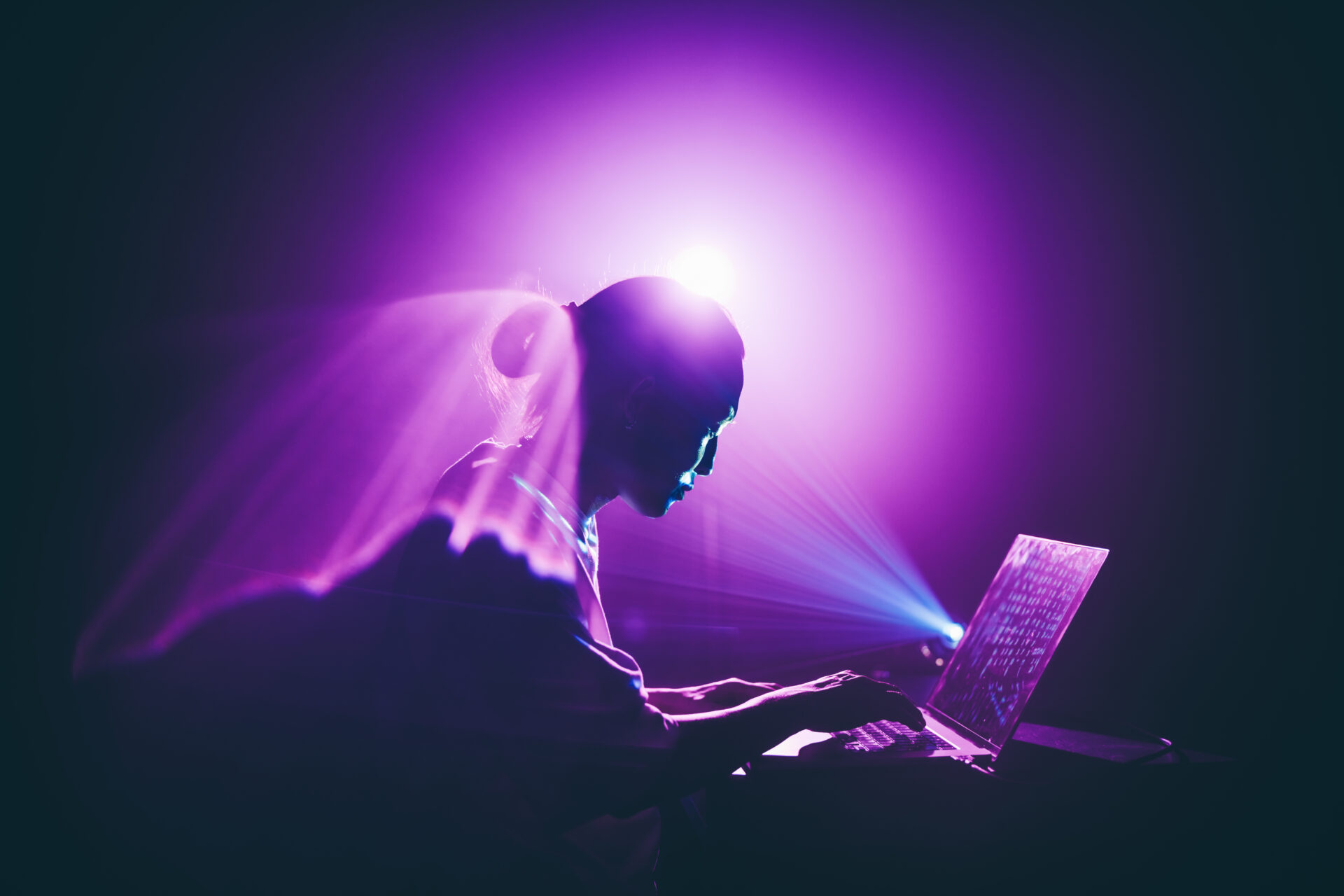 A young person is working on a laptop in a dark, atmospheric room, illuminated by a powerful purple and blue light. The concept of futuristic technology, artificial intelligence, or creative digital work.